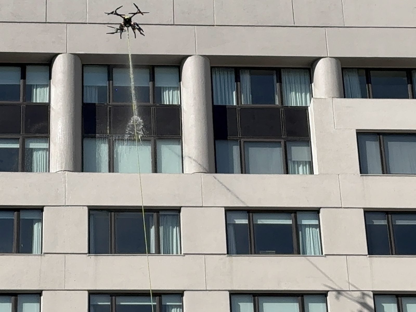Drone de nettoyage relié au sol par un tuyau, pulvérisant de l'eau sur les vitrages d'une façade